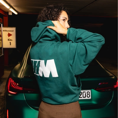 BMW M Pullover in dunkelgrün mit großem M Logo auf dem Rücken, getragen von einem weiblichem Model vor einem BMW M Modell
